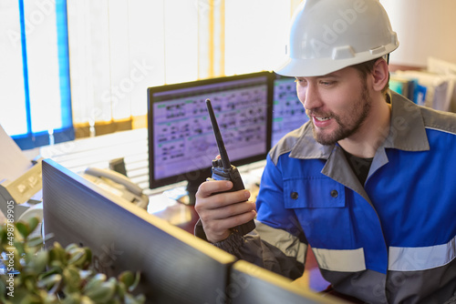 Tableau sur toile Smiling Caucasian worker in overalls and a helmet uses a walkie-talkie, looks at the camera sitting at desk in the control room