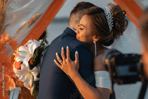 Loving couple at an international wedding at sunset. Beautiful sophisticated bride of Asian appearance. Strong manly groom of Caucasian appearance. Mixed marriages. Warm embrace.