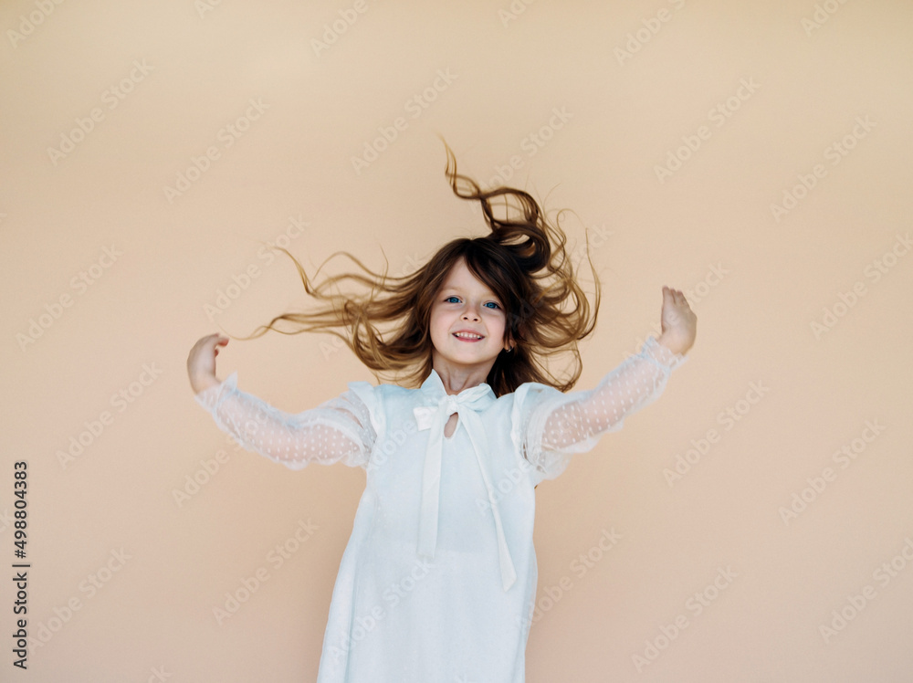 A beautiful little girl in a dress is spinning on a beige background. The concept of a happy childhood, dancing, holidays. Long hair flying, the girl smiles and rejoices. A beautiful little girl in a dress is spinning on a beige background. The concept of a happy childhood, dancing, holidays. Long hair flying, the girl smiles and rejoices.