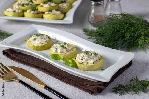 Zucchini slices baked with feta cheese and garlic on a white plate on a gray background