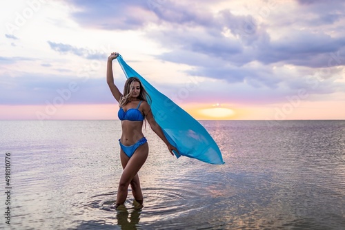 A girl in a blue swimsuit and a bright pareo posing against the background of a sunset in estuary with transparent water