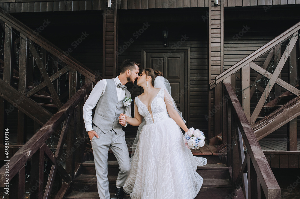 A young, stylish groom in a gray suit and a beautiful bride in a white dress are walking, kissing on a wooden staircase, holding hands, against the backdrop of a large house. Wedding portrait. A young, stylish groom in a gray suit and a beautiful bride in a white dress are walking, kissing on a wooden staircase, holding hands, against the backdrop of a large house. Wedding portrait.