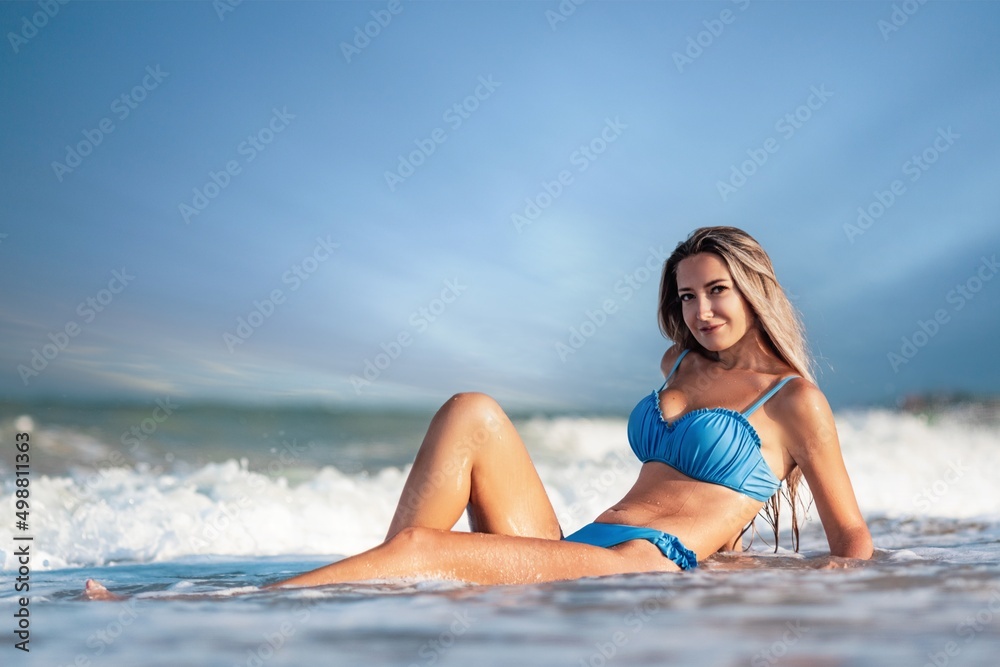 A girl with blond hair in a delicate swimsuit sits by the sea, enjoying the splash of waves