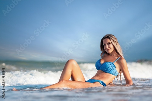 A girl with blond hair in a delicate swimsuit sits by the sea, enjoying the splash of waves