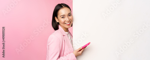 Image of korean female entrepreneur in suit, standing near info wall, advertisement on board, holding smartphone and smiling, posing over pink background