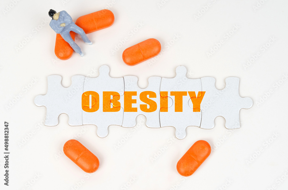 On the white surface of the tablet, a miniature human figure and puzzles with the inscription - OBESITY On the white surface of the tablet, a miniature human figure and puzzles with the inscription - OBESITY