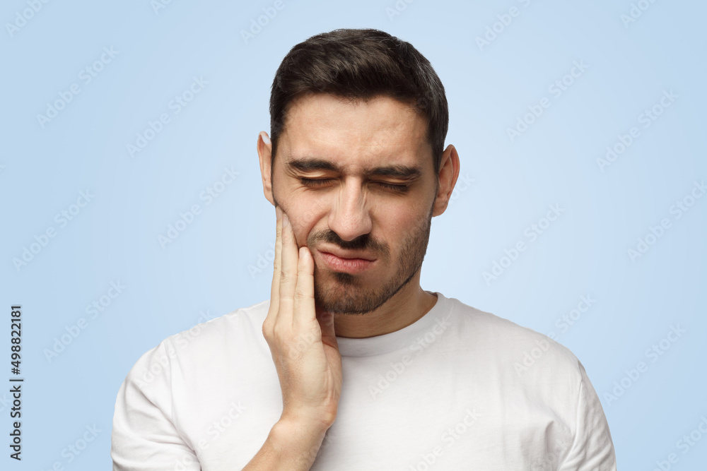 Attractive young man feels pain holding his cheek with hand suffering from bad toothache