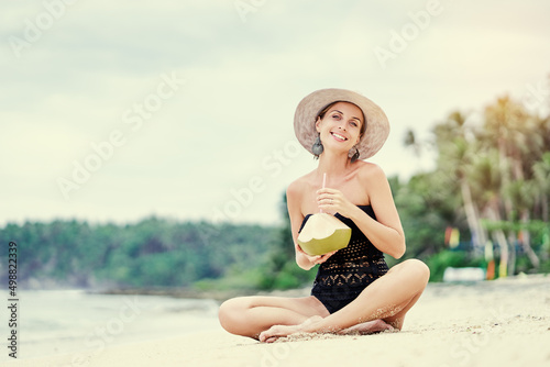 Vacation on the seashore.Young woman in hat and swimsuit holding green cococnut relaxing on the white sand beach.