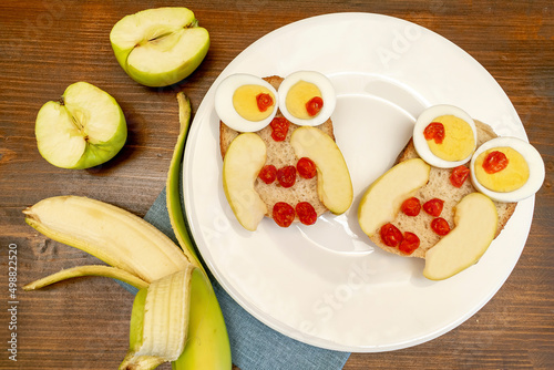 Funny owl face sandwich toast bread with chicken eggs, apple, banana, dried berry fruits on plate. Cute kids childrens baby's sweet dessert breakfast,lunch, food art on wooden background,top view