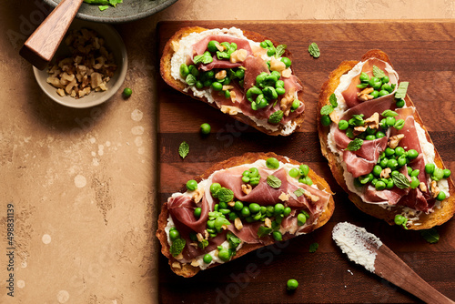 A pea, prosciutto, and ricotta crostini with a bite missing on a plate next to a bowl of peas, walnuts, and flakey sea salt. photo