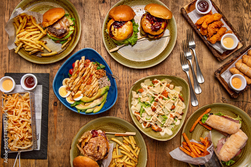 Set of fast food dishes on wooden table with assorted burgers, sandwiches, sweet potato fries, tequeños, fried chicken strips and caesar salad, chicken salad with avocado and boiled egg