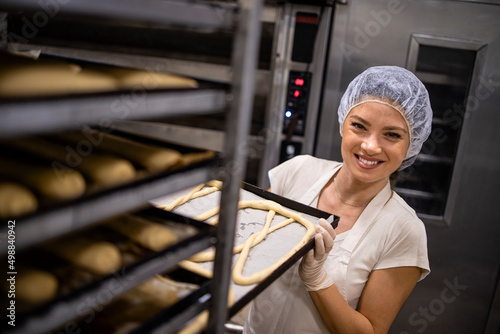 Portrait of female baker in white uniform and hairnet working in bakery production moving tray with raw bread and pastries. photo