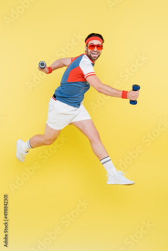 Smiling sportsman in sunglasses jumping and holding dumbbells isolated on yellow.