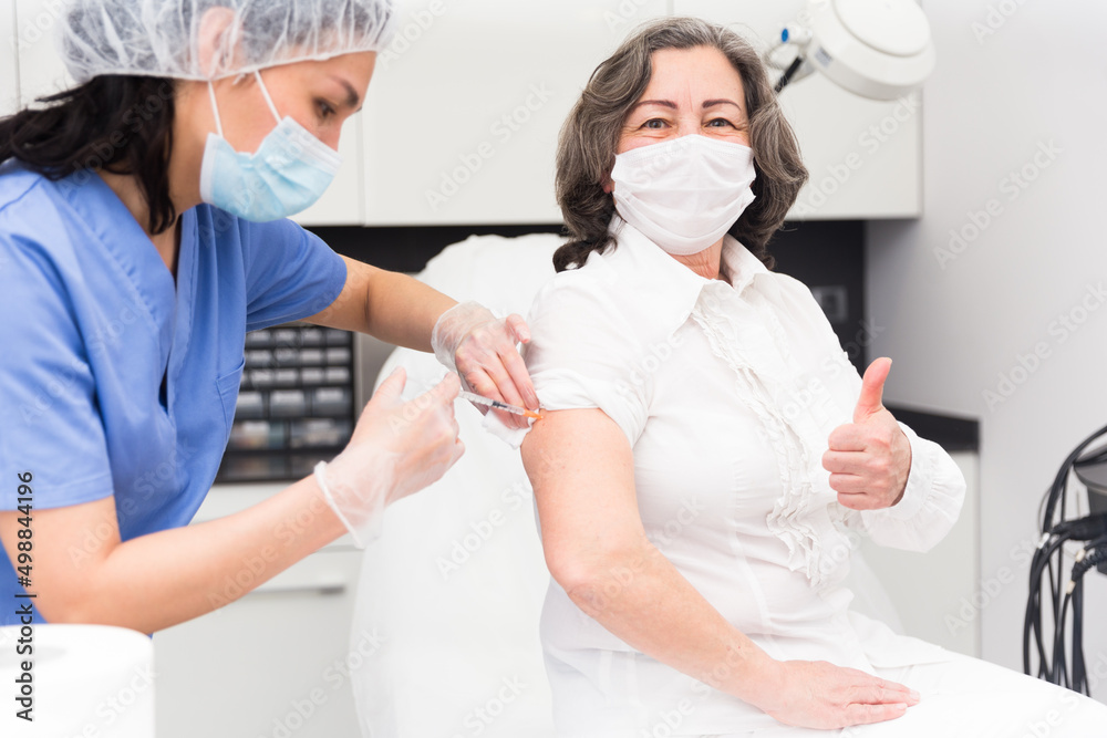 Nurse in protective mask making injection of vaccine to mature woman