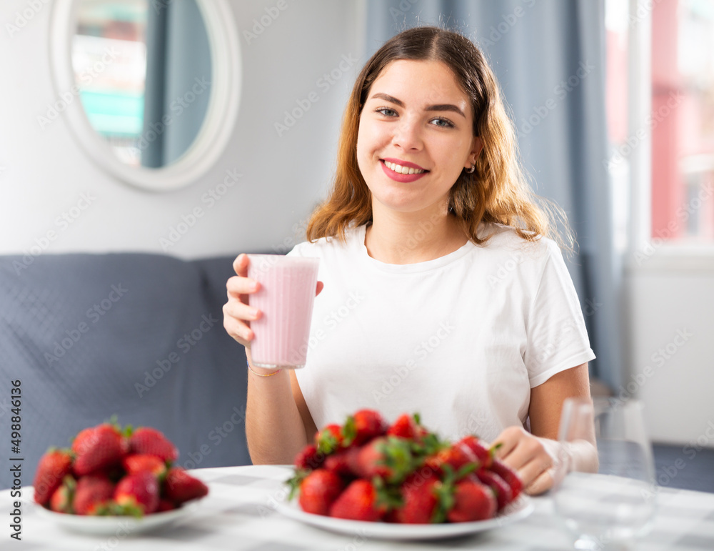 Portrait of a happy young woman holding a strawberry smoothie in her hand and ripe strawberries on the table at home