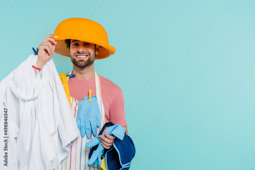 Cheerful man in clothespins holding wash bowl on head and clothes isolated on blue. Cheerful man in clothespins holding wash bowl on head and clothes isolated on blue.