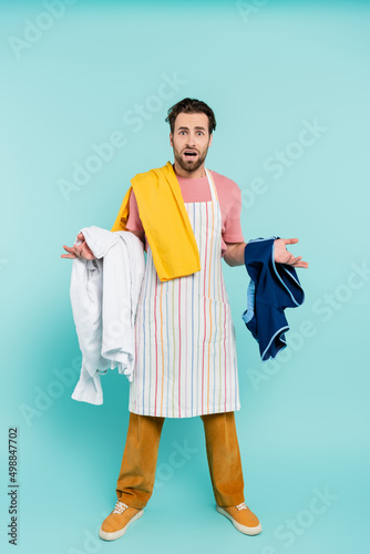 Confused unmarried man in apron holding clothes on blue background.