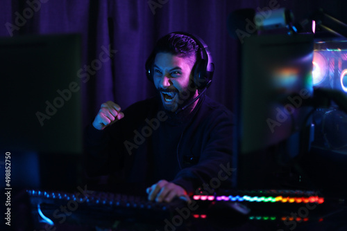 A young man plays a computer game in the midst of colorful neon lights. The gameplay is fun and winning. Smiling and raising fists with satisfaction in the black studio. vertical photo