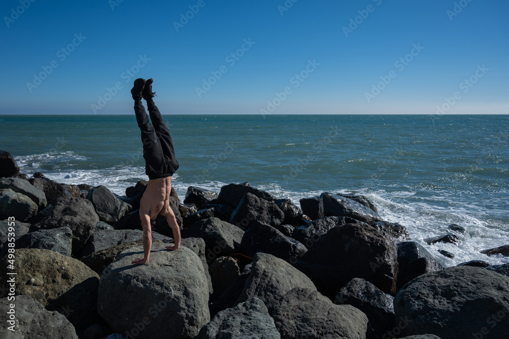 Shirtless man doing handstand on rocks by the sea. 