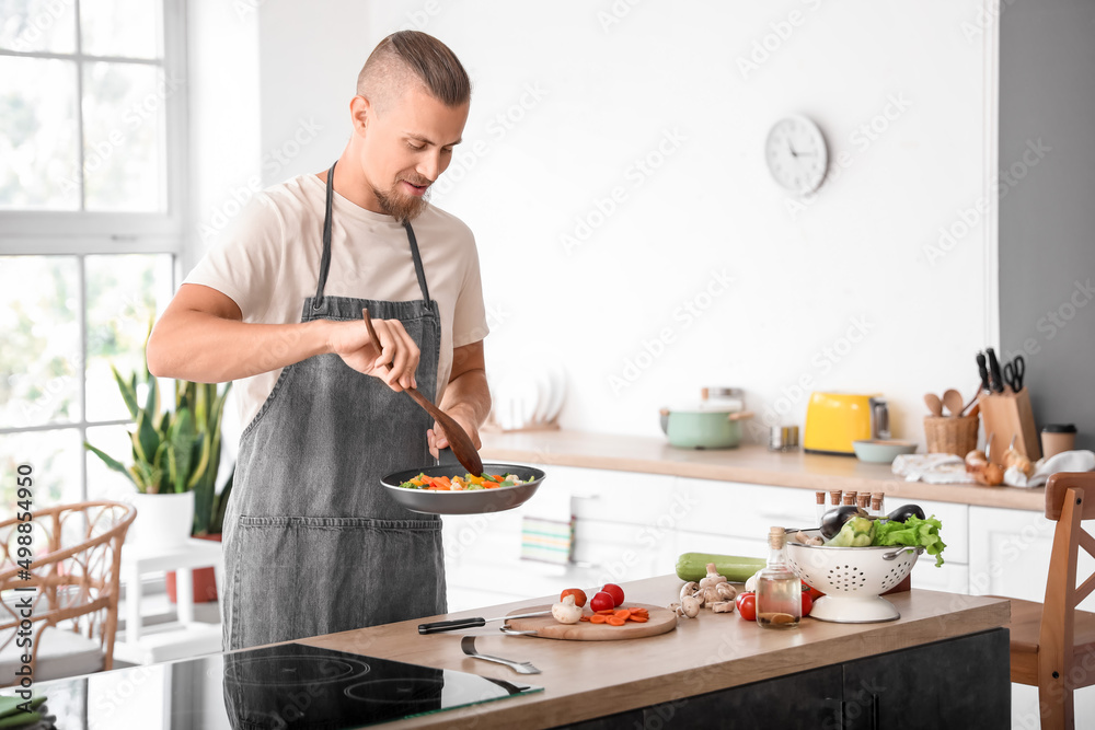 Handsome young man with spatula frying vegetables in kitchen