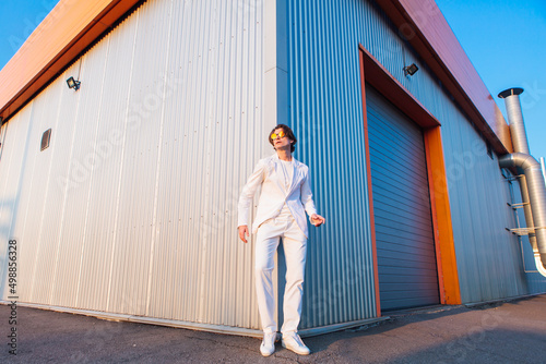 Wide angle portrait of a handsome man dressed in a white suit and round hippie sunglasses standing near the corner of a building during sunset