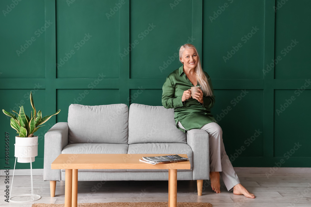 Obraz premium Mature woman drinking tea while sitting on sofa near green wall