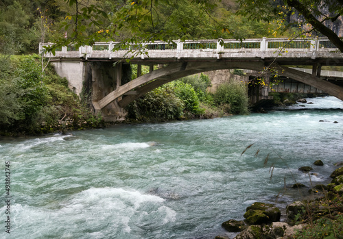 A mountain river with bubbling turquoise water flows in the picturesque mountains of Abkhazia. The old transport bridge hangs in an arc over the river, along the banks of which stones are scattered