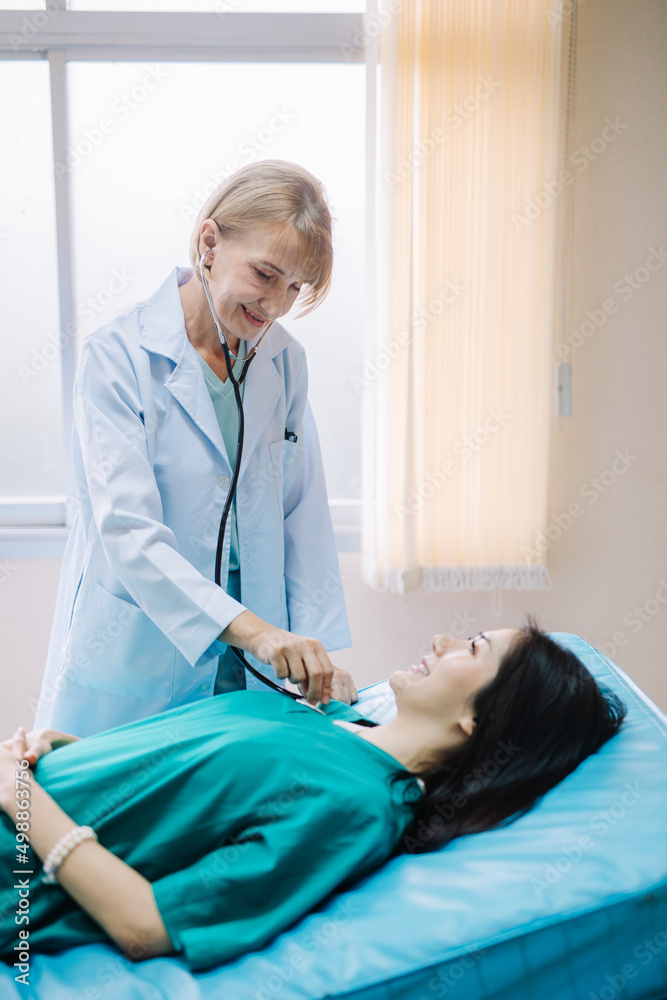 Fototapeta premium A female doctor examining a patient with a stethoscope while smiling at the patient lying on the bed