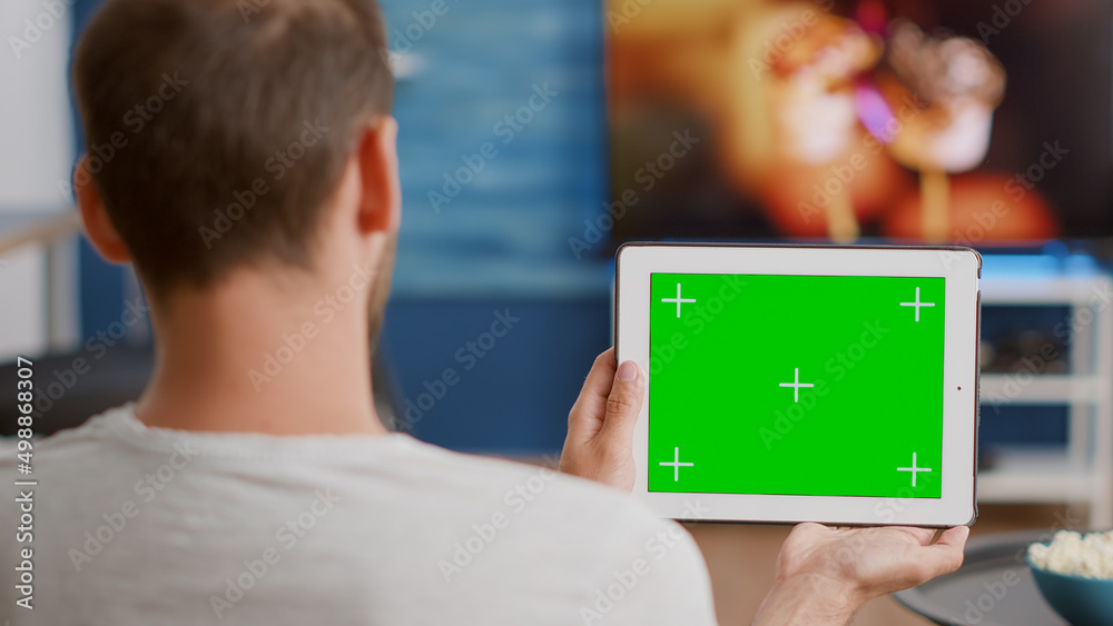Closeup of man holding digital tablet with green screen watching online video content sitting on sofa in front of tv. Person looking at touchscreen device with chroma key enjoying influencer vlog. Closeup of man holding digital tablet with green screen watching online video content sitting on sofa in front of tv. Person looking at touchscreen device with chroma key enjoying influencer vlog.