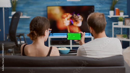 Static tripod shot of man holding smartphone with green screen watching online video content with girlfriend sitting on sofa. Couple enjoying social media videos on mobile phone with chroma key .
