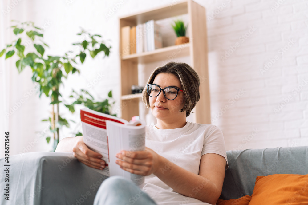 Girl with glasses reads book on the couch in front of the bookshelf.