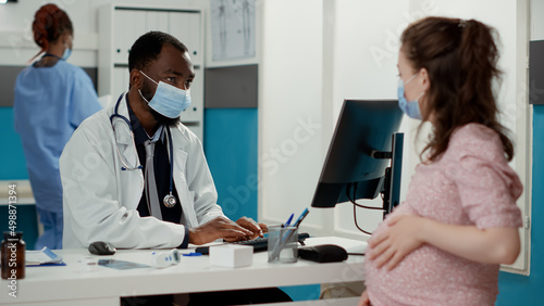 General practitioner examining woman with pregnancy belly during coronavirus pandemic. Male specialist talking to patient expecting child, giving medical advice and helping with maternity.