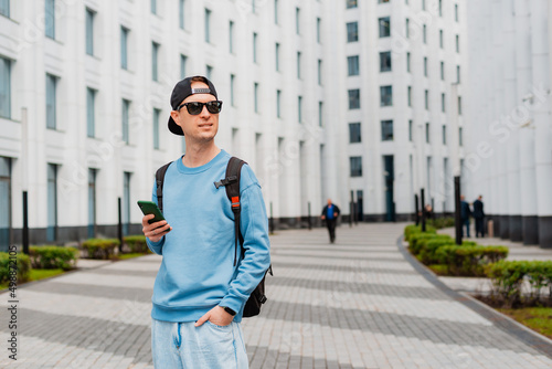 Positive hipster guy with backpack standing outdoor holding smartphone while looking away with smile