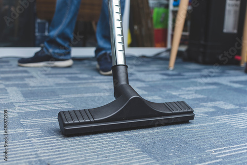 A man cleans the carpet flooring of an office with a vacuum cleaner with an attached floor sweeper nozzle.