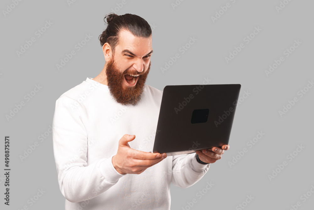 Bearded male hipster is holding an open laptop and screaming at it over grey background.