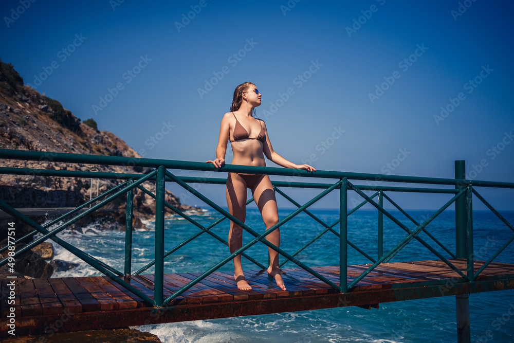 beautiful young woman in a brown swimsuit walks on the pier by the beach on the sea on a summer sunny day