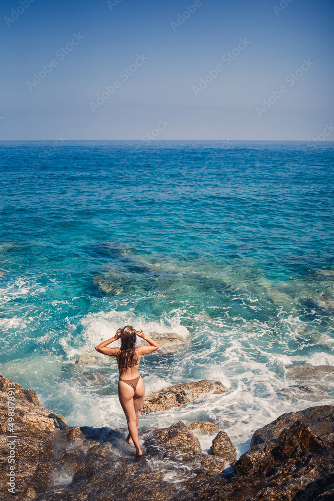 Sexy woman in full body swimsuit with long hair walks on large rocks on a rocky beach during a storm at sea. Back view. Woman swimwear island tropics landscape exotic walk Sexy woman in full body swimsuit with long hair walks on large rocks on a rocky beach during a storm at sea. Back view. Woman swimwear island tropics landscape exotic walk