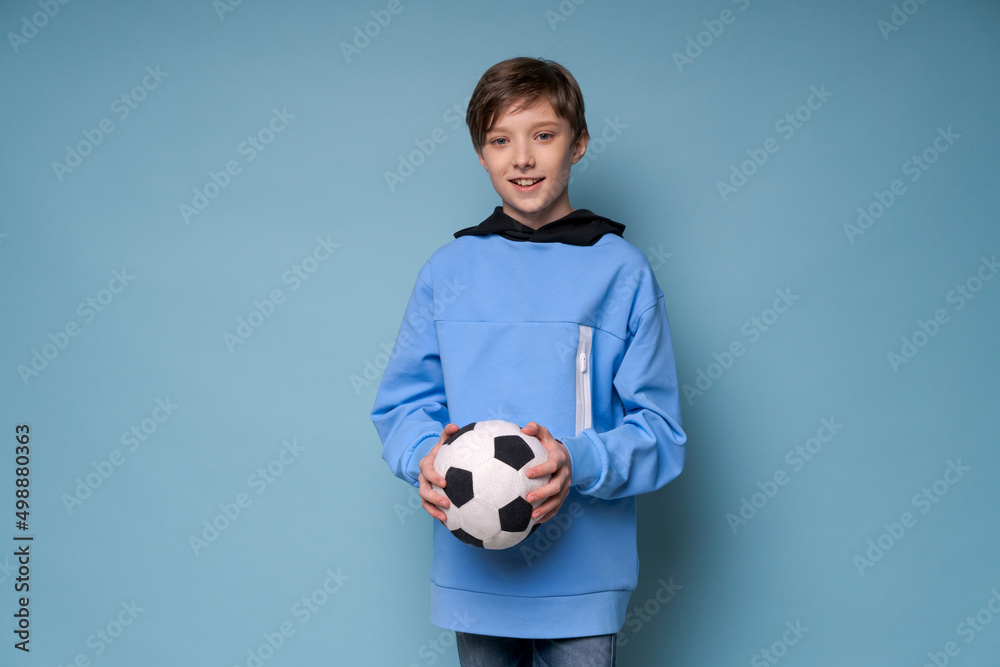 Fan sports boy teenager player holding soccer ball celebrating happy smiling laughing free text copy space copy space on colored background in blue sweatshirt posing cheerful guy
