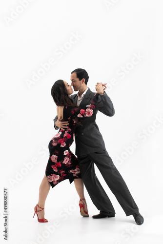 Couple of professional tango dancers in elegant suit and flowery print dress pose in a dancing movement on white background. Handsome man and woman dance looking eye to eye.