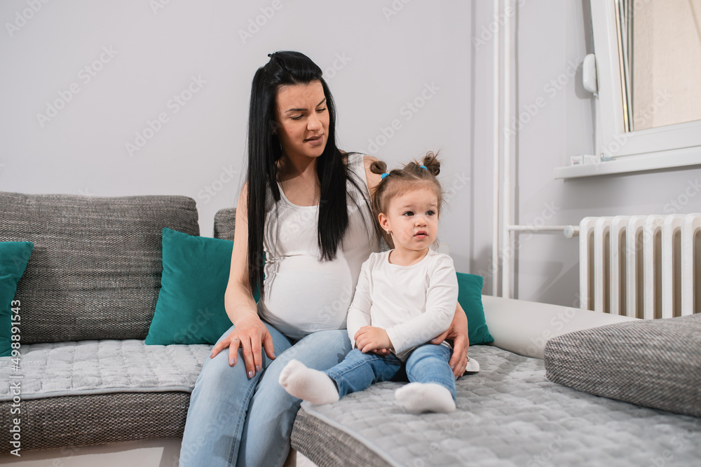 Proud mom looks at her beautiful daughter with ponytails while little cute kid looks aside. Two caucasian females sit on the sofa in a sitting room close to each other. Proud mom looks at her beautiful daughter with ponytails while little cute kid looks aside. Two caucasian females sit on the sofa in a sitting room close to each other.
