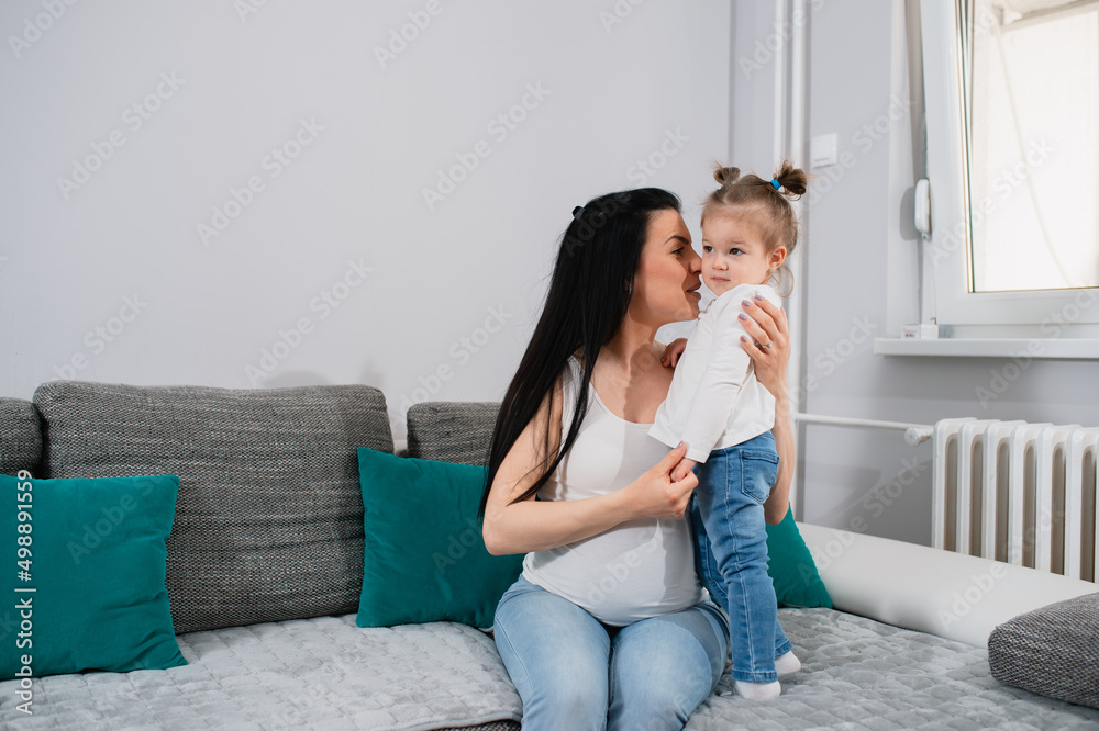 Pregnant mother and her daughter caress on sofa in living room. Beautiful girl stands on a couch while her caucasian mom sits and hugs her with left hand and holds kid's hand. A little girl looks away Pregnant mother and her daughter caress on sofa in living room. Beautiful girl stands on a couch while her caucasian mom sits and hugs her with left hand and holds kid's hand. A little girl looks away