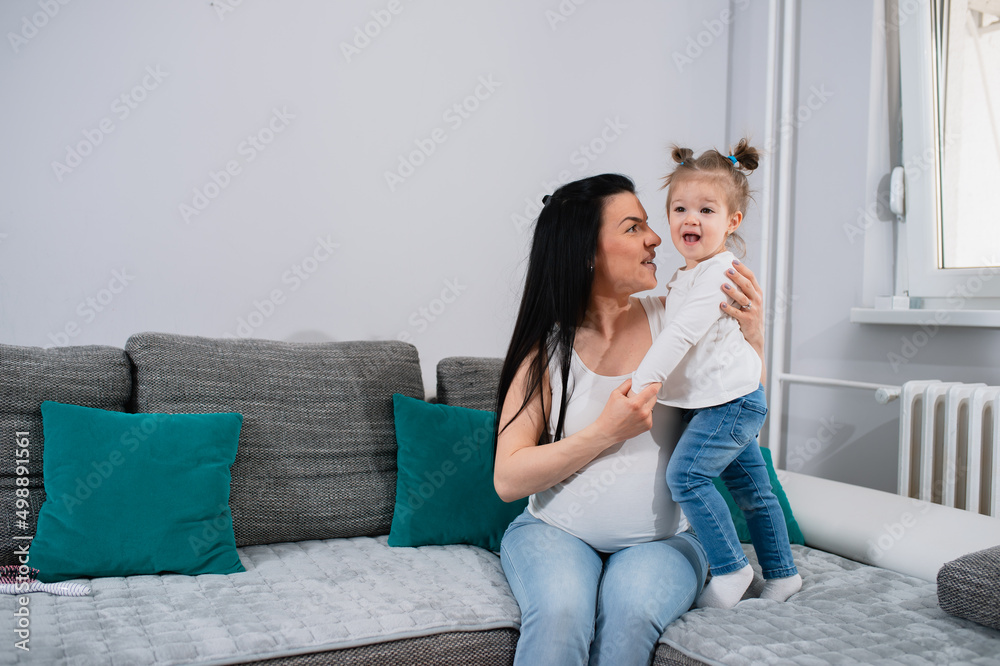 The pregnant mother and daughter spend a wonderful time in the living room. Little lady with ponytails stands on the sofa beside her mom who sits.Caucasian woman hugs her first child and looks at her.