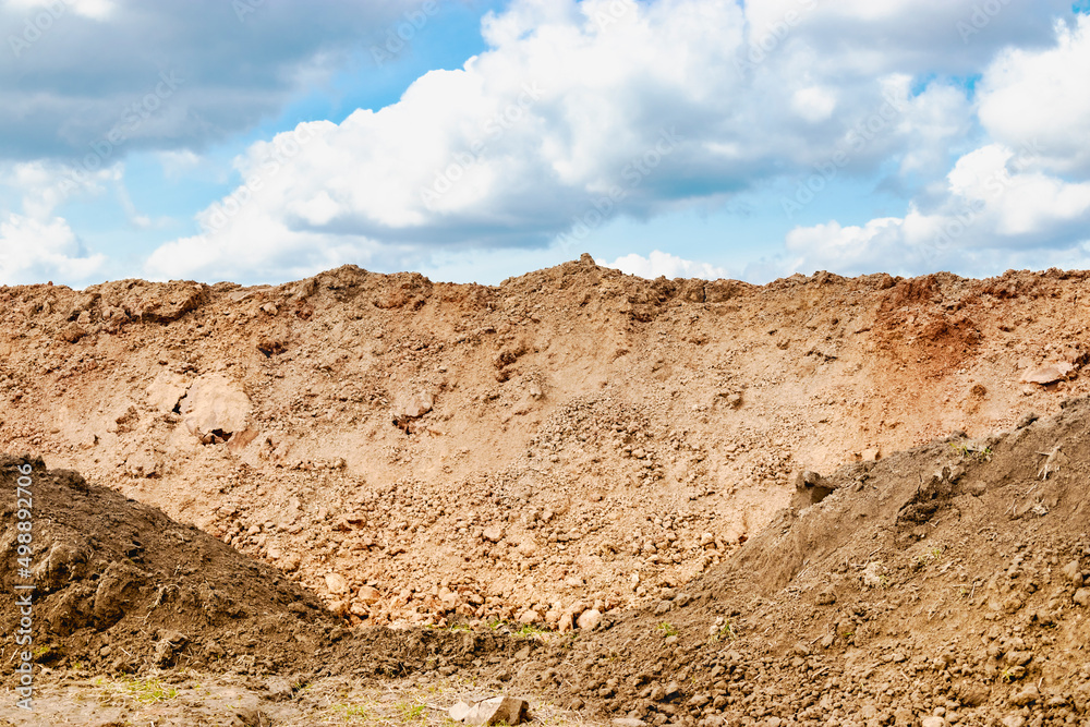 A pile of soil dug out of a trench against a blue cloudy sky. Earthworks at the construction site.