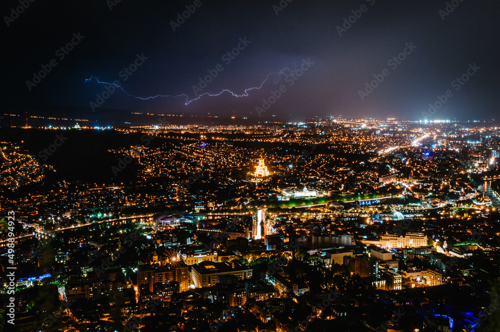 View from Mtatsminda mountain of night sky with lightning, a thunderstorm over new and old houses, tower, city, town, and streets in Tbilisi, is the capital of Georgia.