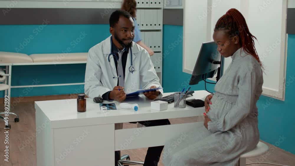 African american doctor and patient expecting child meeting in cabinet at checkup visit. Male physician and pregnant woman having conversation about childbirth at consultation appointment. African american doctor and patient expecting child meeting in cabinet at checkup visit. Male physician and pregnant woman having conversation about childbirth at consultation appointment.