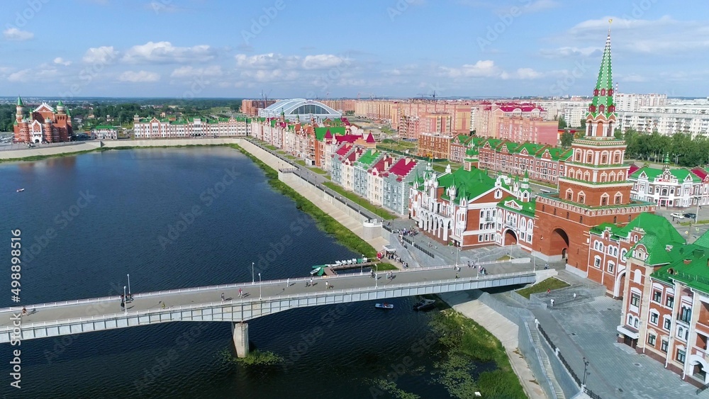 The promenade of a beautiful city on a summer day. A drone flies over a pedestrian bridge over the water. People walk along the pavement along the low brick buildings. Catamarans float along the river