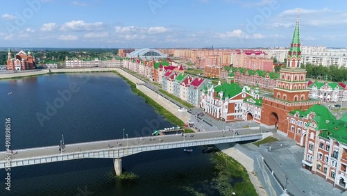 The promenade of a beautiful city on a summer day. A drone flies over a pedestrian bridge over the water. People walk along the pavement along the low brick buildings. Catamarans float along the river