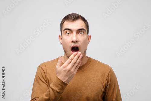 portrait of surprised caucasian brunette man with hand covering his open mouth in surprise and big dumbfounded eyes on grey background