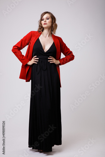 High fashion photo of a beautiful elegant young woman in a pretty black evening party dress with a deep neckline, red jacket posing on white background. Slim figure, hairstyle, studio shot.