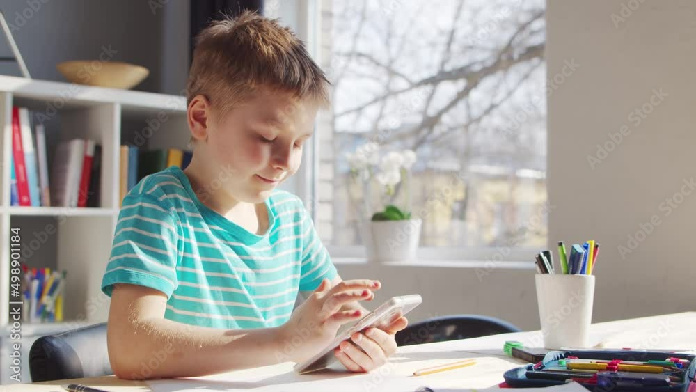 Boy is Doing his Homework at the Table. Cute Child is Learning at Home with the Help of Smartphone device and School Education Textbooks. Study and Entertainment Concept . Boy is Doing his Homework at the Table. Cute Child is Learning at Home with the Help of Smartphone device and School Education Textbooks. Study and Entertainment Concept .